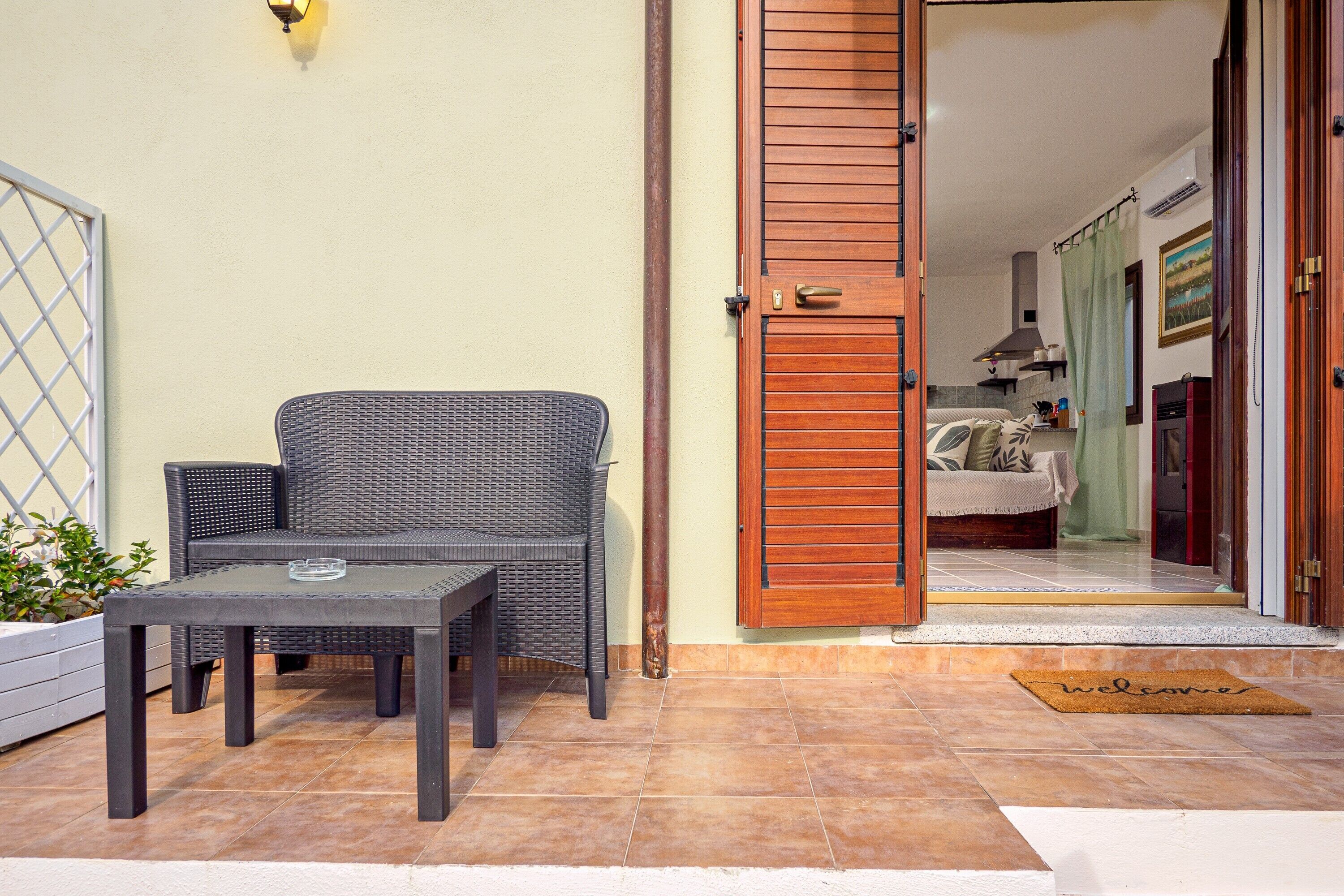 Un espace extérieur aménagé avec une table et des chaises, une porte en bois donnant sur une pièce, et un paillasson.