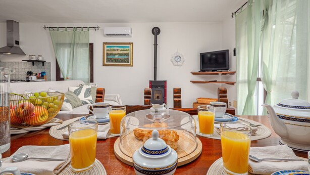 Une table mise pour le petit-déjeuner, avec une nappe, de l’assiette, des verres, une théière et un panier de fruits.