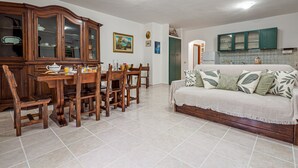 Un coin repas avec une table et des chaises en bois, un meuble avec des portes en verre, un canapé agrémenté de coussins à motifs et un espace cuisine avec des meubles verts.
