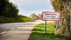 Una señal de tráfico que indica 'PRAPOUTEL LES 7 LAUX comuna de LES ADRETS', con una carretera y vegetación al fondo.