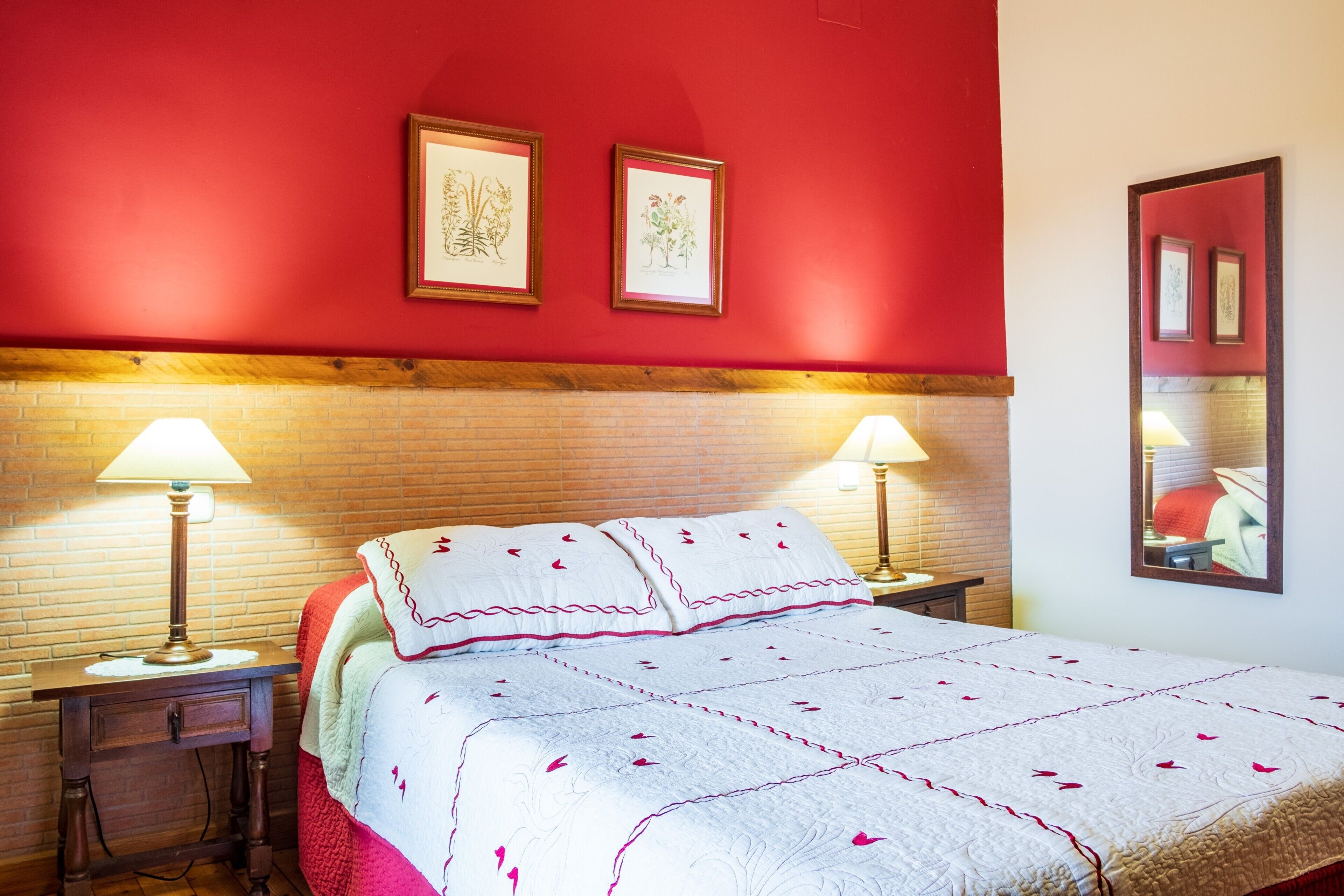 Habitación de hotel con pared roja, cabecera de madera, cama con ropa de cama blanca y detalles rojos, dos lámparas de noche y cuadros en la pared.