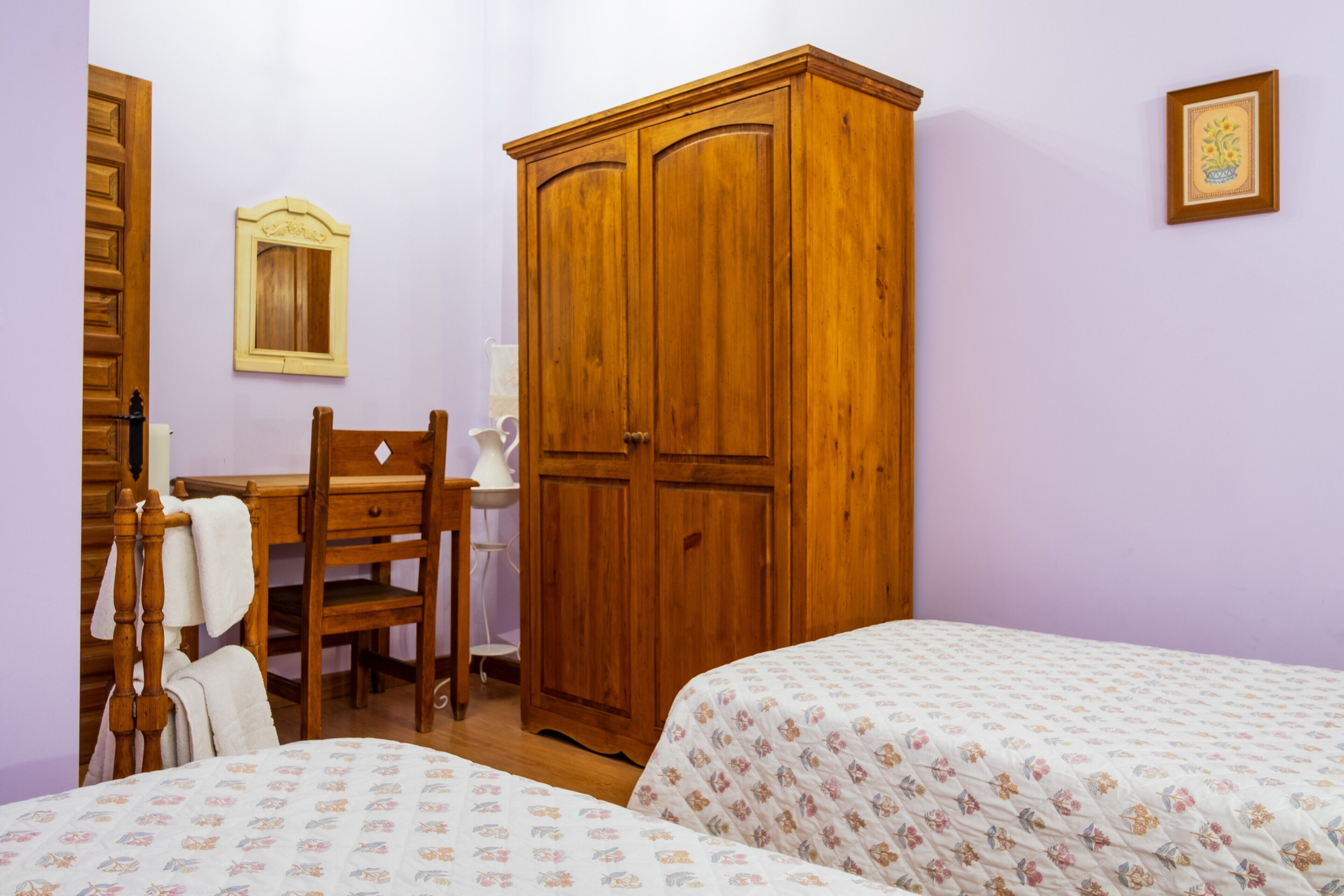 A bedroom with two beds, a wooden wardrobe, a desk with a chair, and a small bedside table.