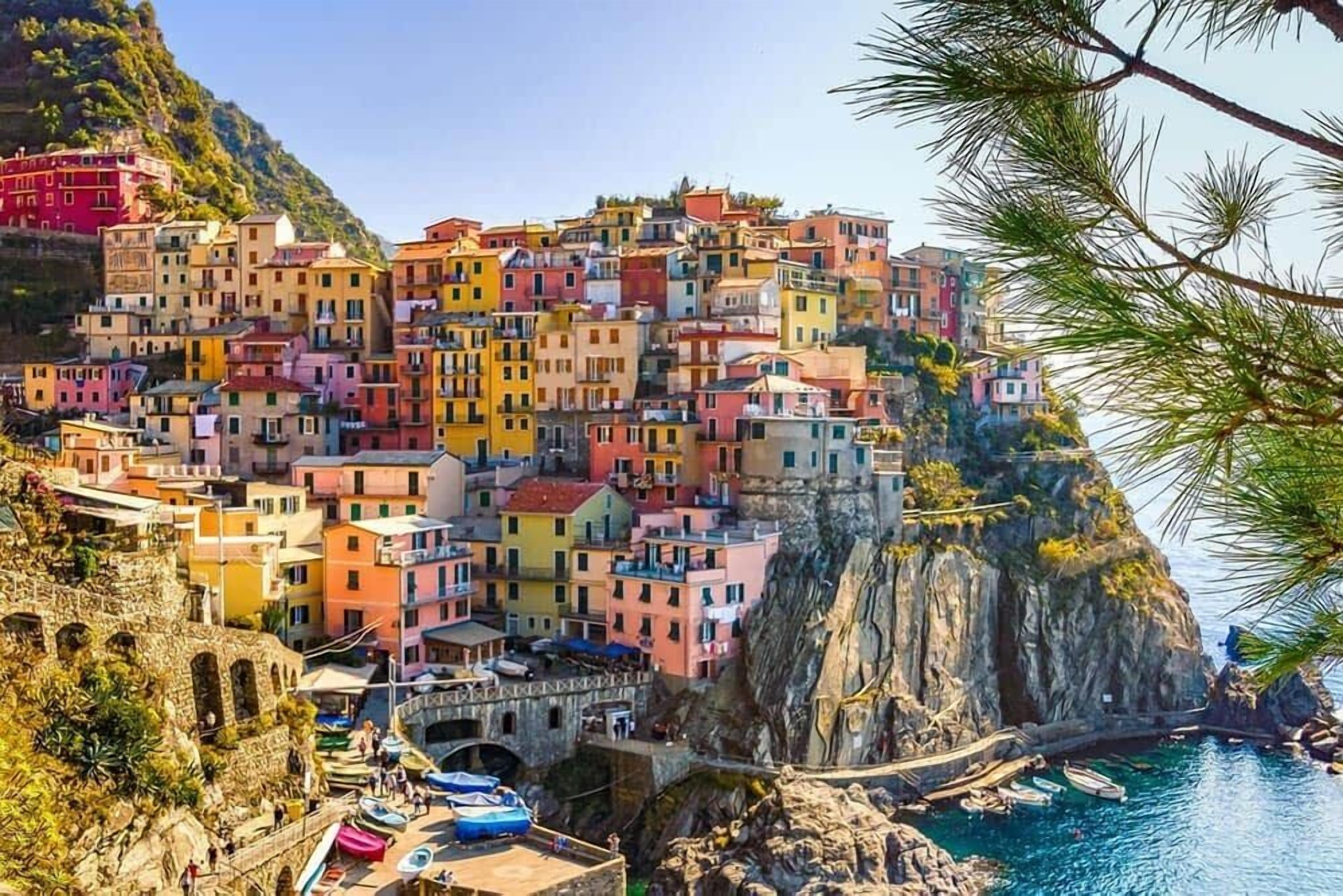 A coastal village with colorful buildings perched on a cliff, a stone bridge, and boats at the harbor.