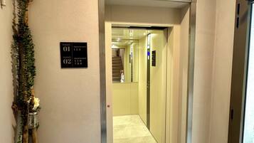 Een interieurhal met een lift, een bord waarop 'PIANO 01' en 'PIANO 02' appartementen staan, en een sierplant met witte bloemen.