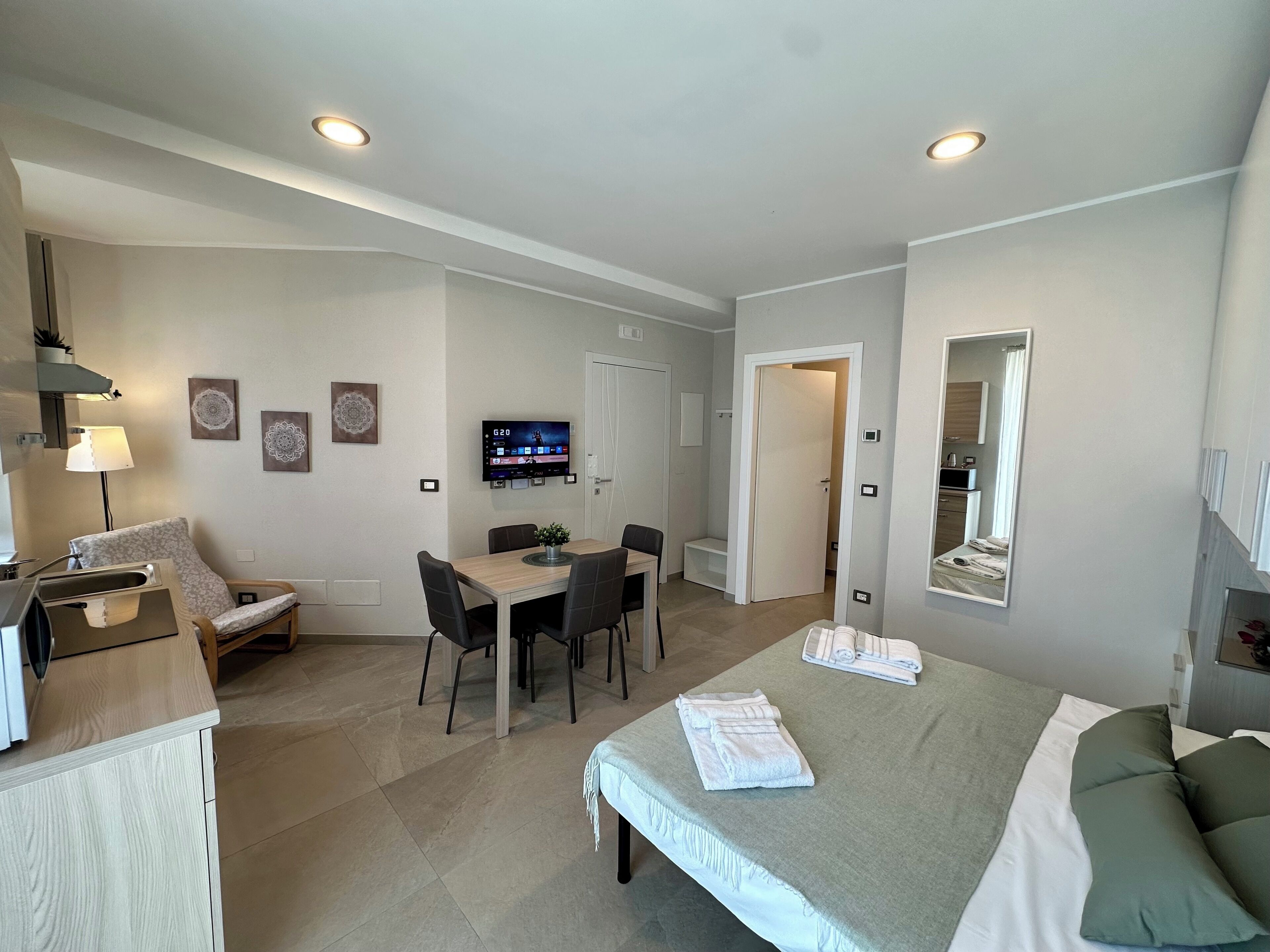 A modern hotel room with a bed, a desk with a chair, a dining table with chairs, a TV mounted on the wall, and a bathroom visible through an open door.