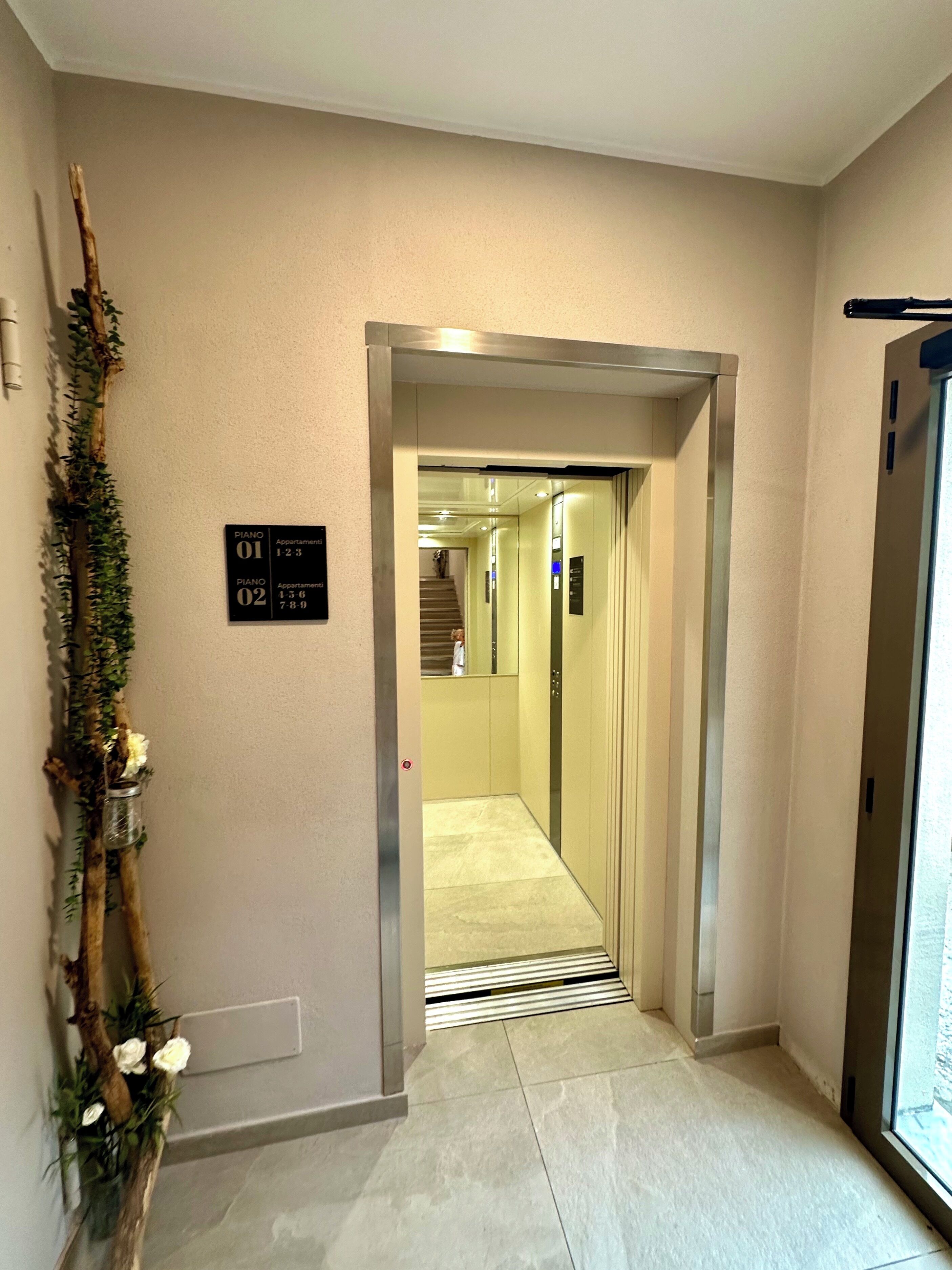 An interior hallway with an elevator, a sign indicating 'PIANO 01' and 'PIANO 02' apartments, and a decorative plant with white flowers.