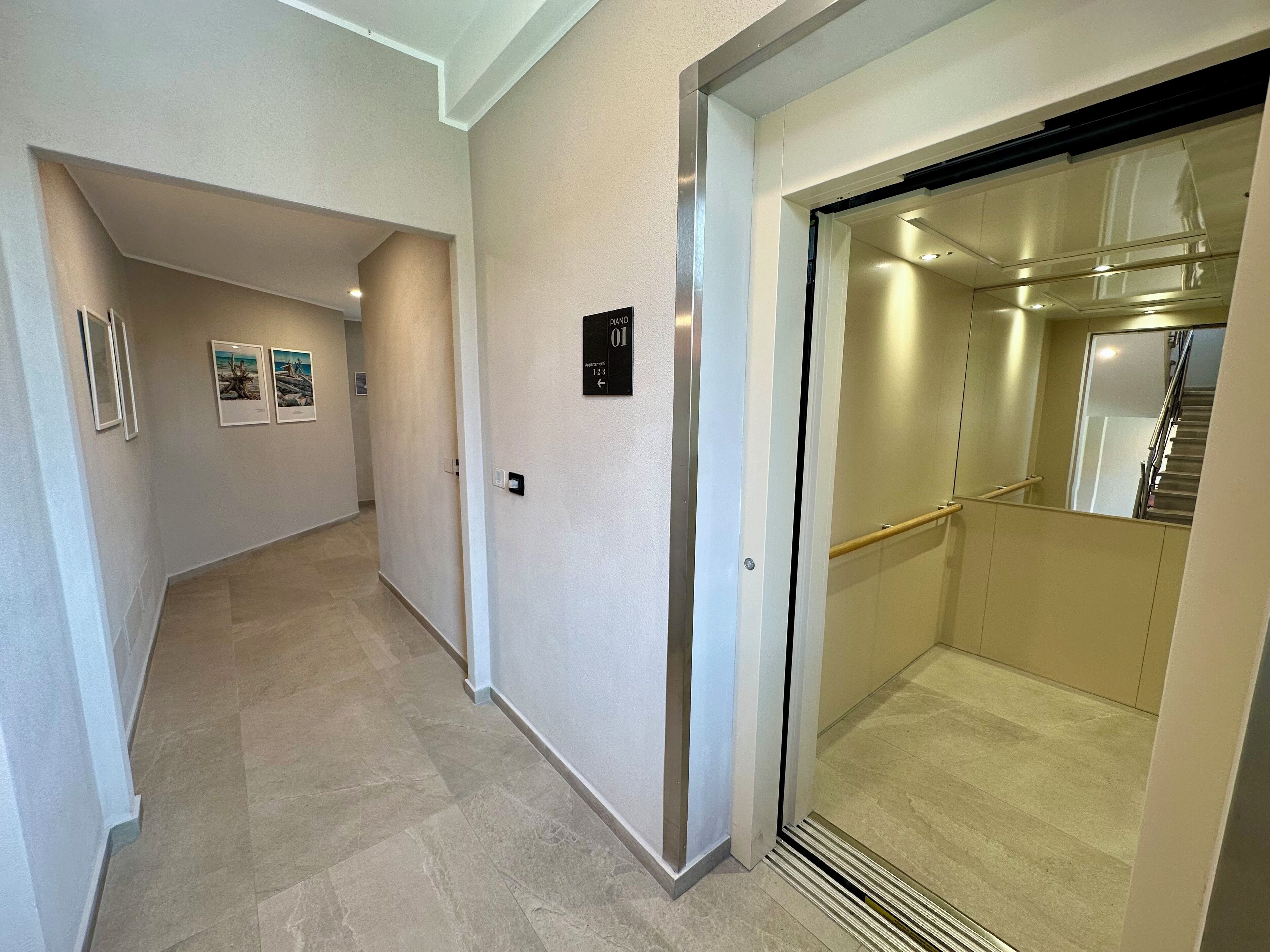 Un corridoio con una porta che conduce a una scala e ad un ascensore. Alle pareti sono presenti quadri incorniciati e un segnale che indica la posizione dell'ascensore.
