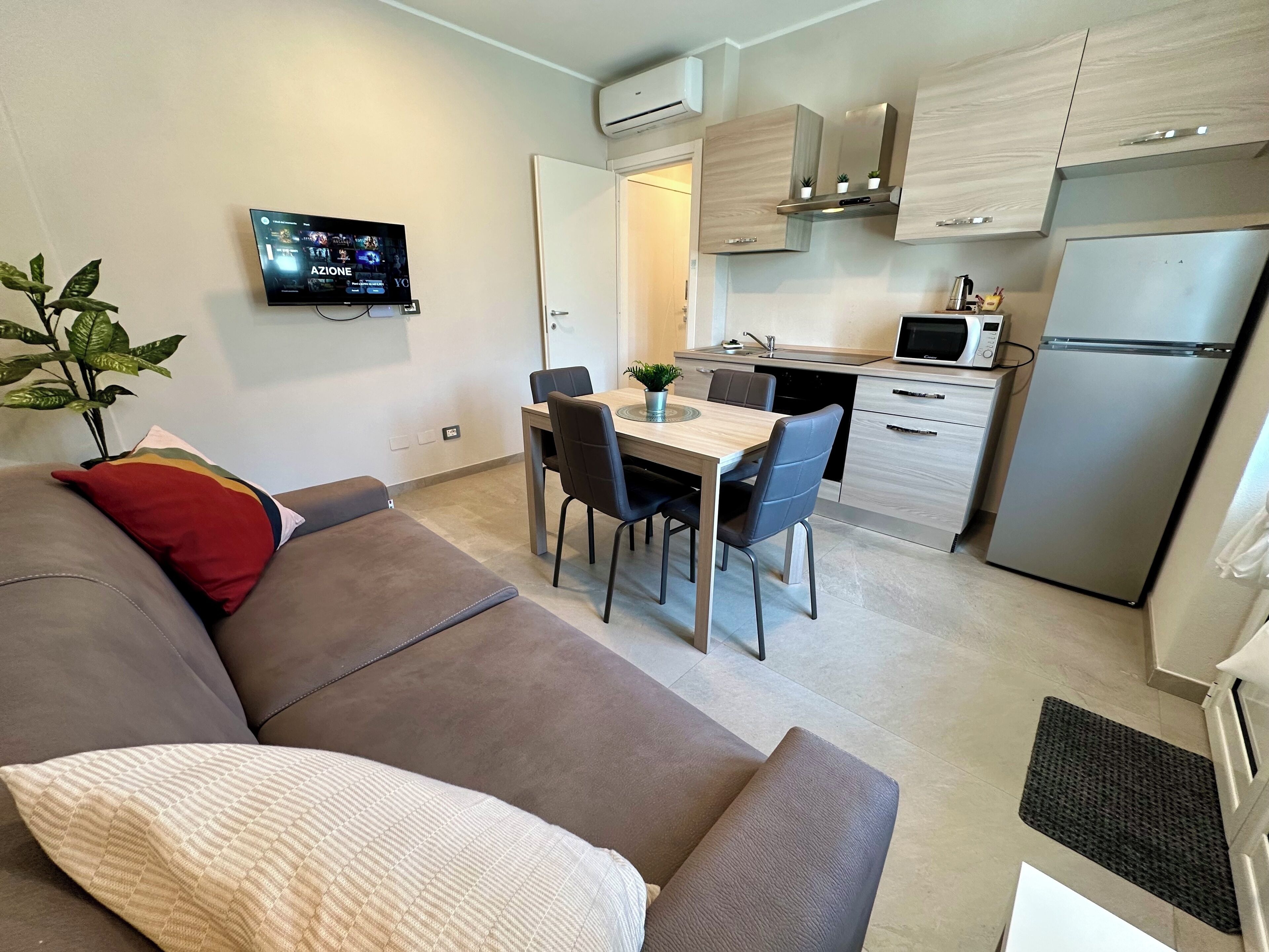 Un salon moderne avec un canapé gris, une petite table à manger avec des chaises, un téléviseur fixé au mur et un coin cuisine équipé d’un réfrigérateur, d’un four à micro-ondes et de placards.
