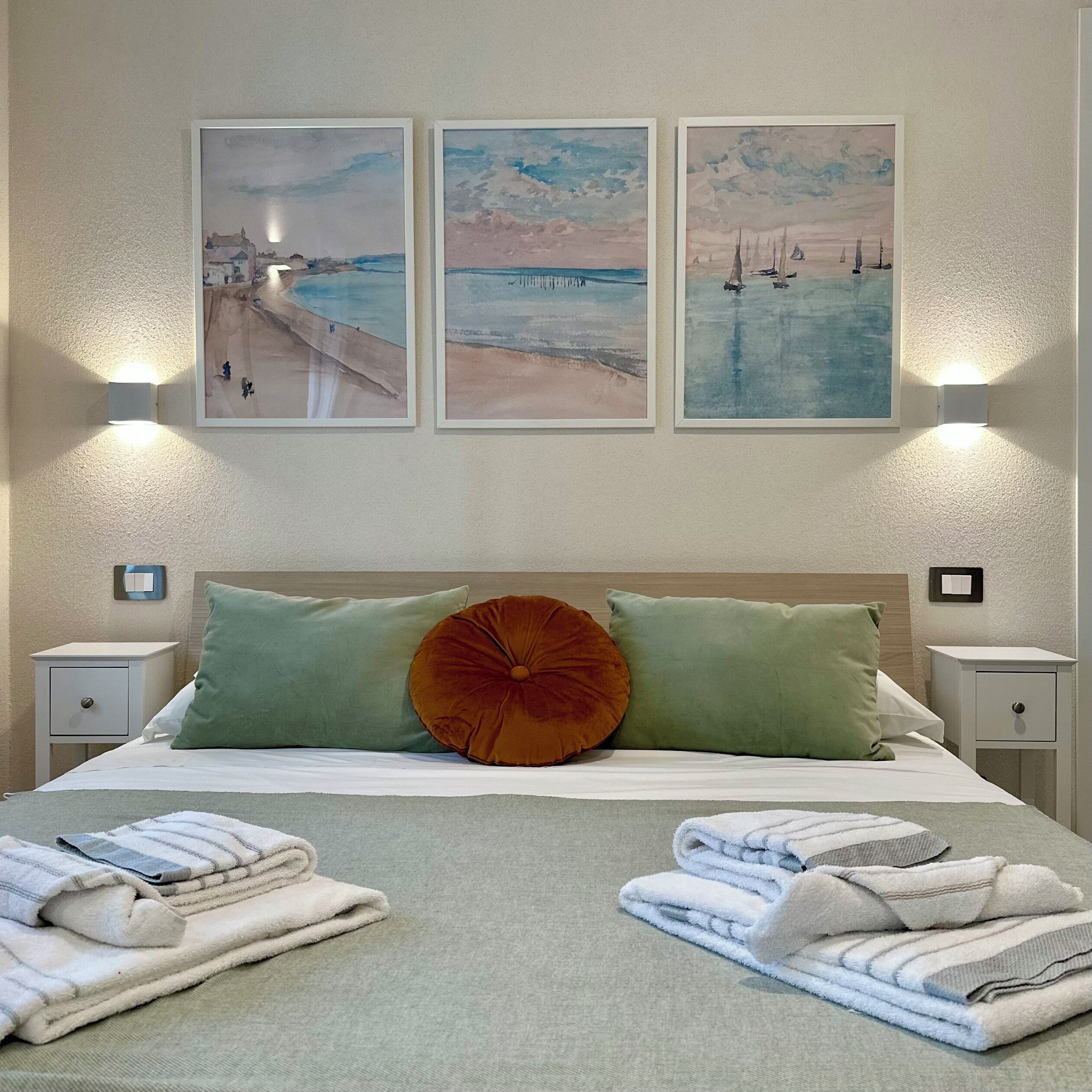 Habitación de hotel con una cama, almohadas verdes y naranjas, y tres cuadros en la pared.