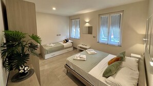 A modern bedroom with two single beds, a large window with curtains, a bedside table, a mirror, and a potted plant.