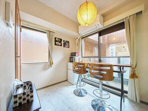 A hotel room with a dining table, two chairs, a hanging light fixture, an air conditioner, a window with curtains, and a small kitchenette area.
