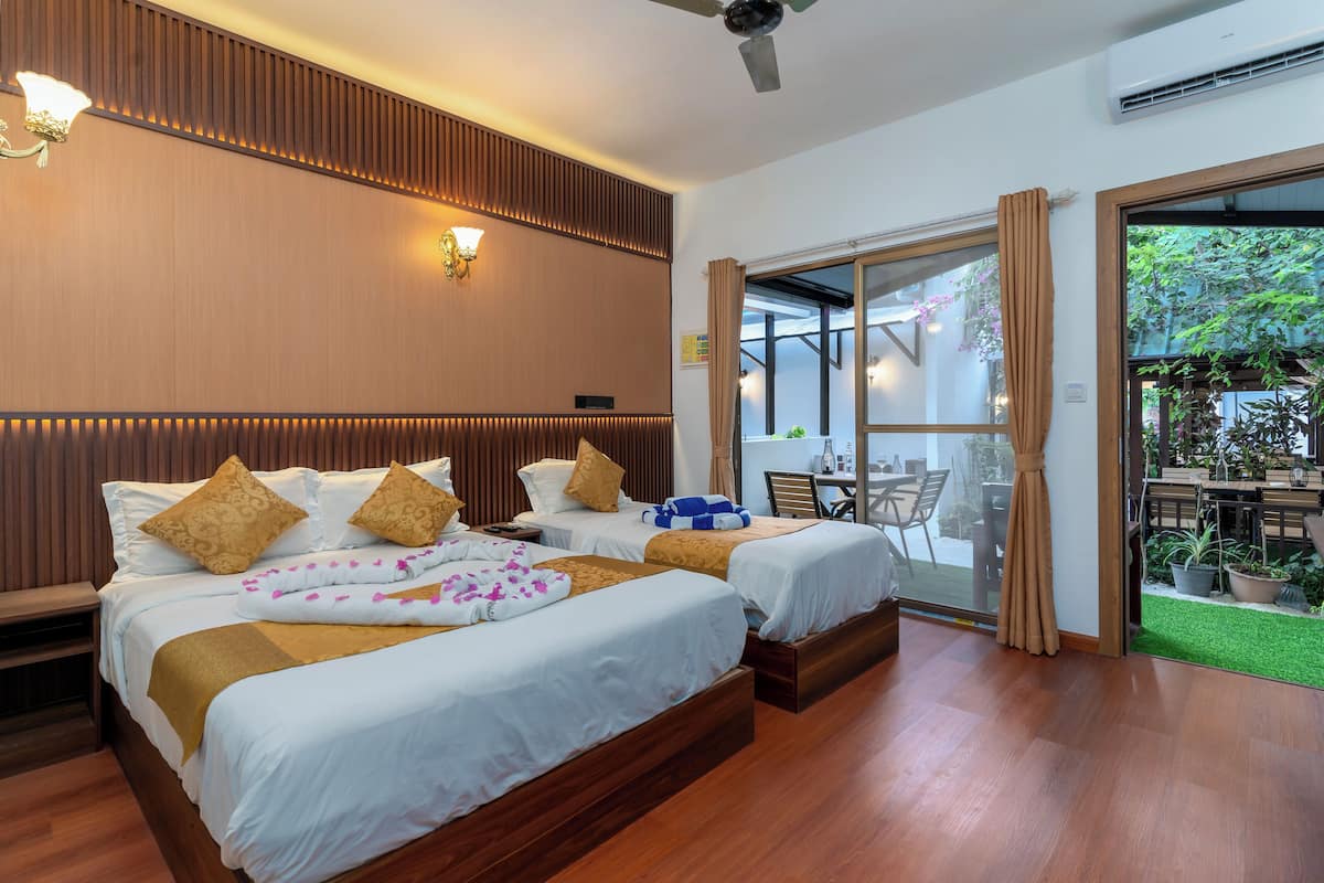 A hotel room with two beds, wooden paneling, a ceiling fan, and a sliding glass door leading to an outdoor area with plants.