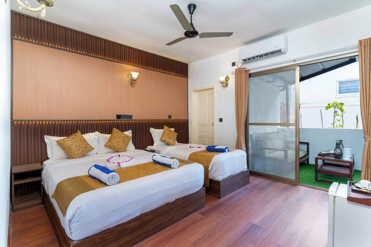 A hotel room with two beds, a ceiling fan, a sliding glass door to a balcony, and a small table with a vase.