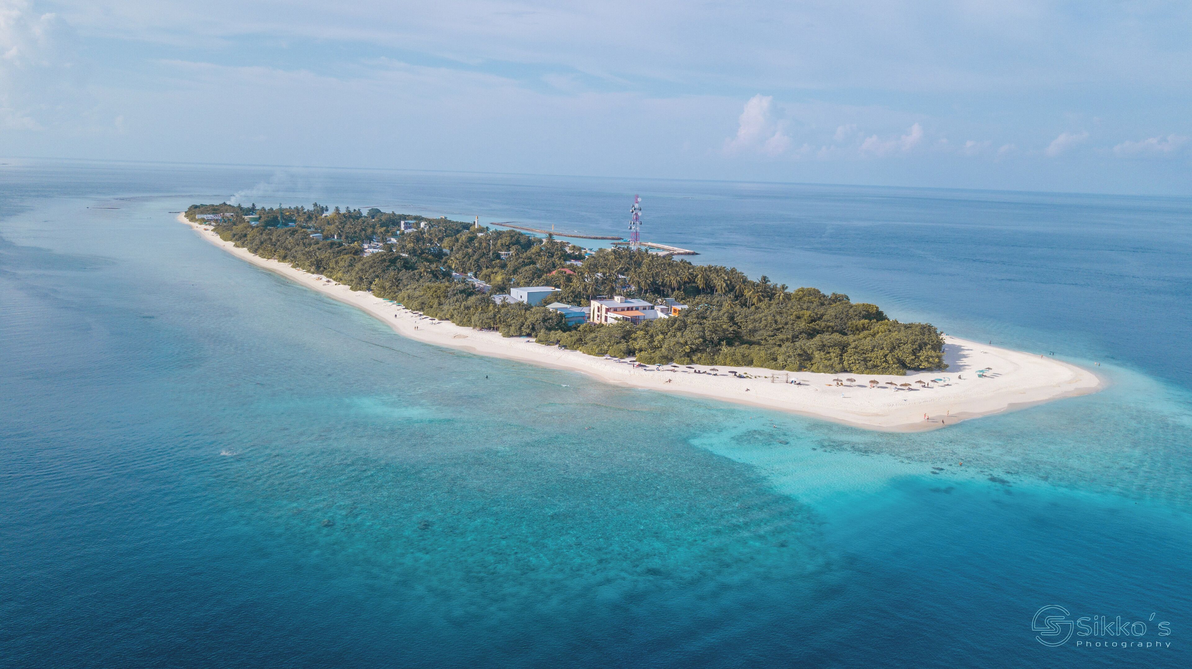 An aerial view of a tropical island with a long sandy beach, dense greenery, and a few buildings.