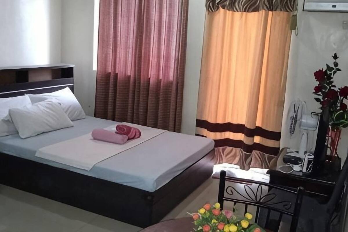 A hotel room with a bed, curtains, a bedside table with a lamp, and a vase with flowers.