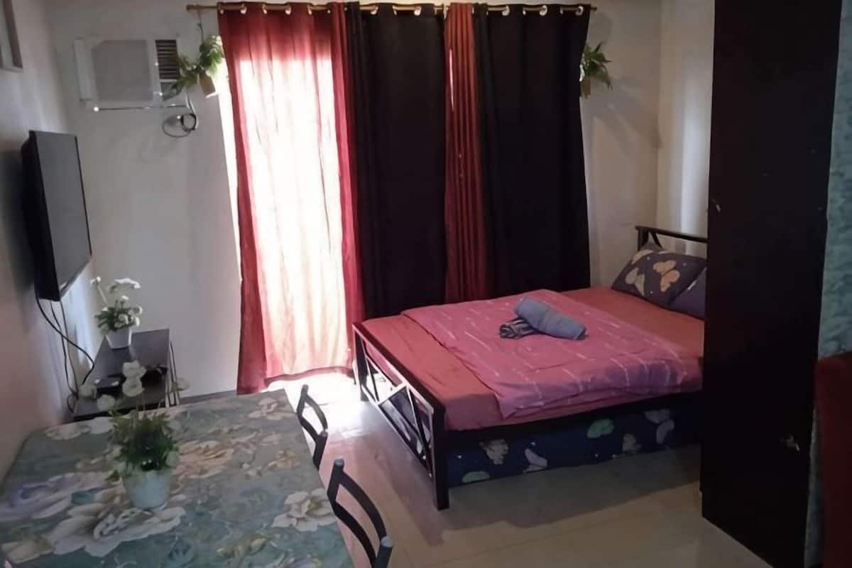 A bedroom with a bed, curtains, a TV, a desk with a chair, and a window.