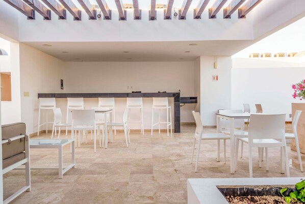 Een modern buitenzitgedeelte met witte stoelen en tafels, een centrale bar en een pergola erboven.