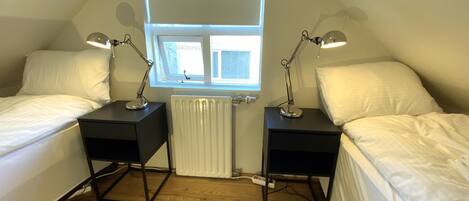 A small, well-lit room with two single beds, a window with a 'No Smoking' sign, and a radiator.