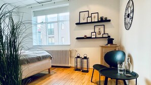 Un dormitorio moderno con cama, radiador, ventana, planta, mesa con vasos y una botella, y estantes con fotos.