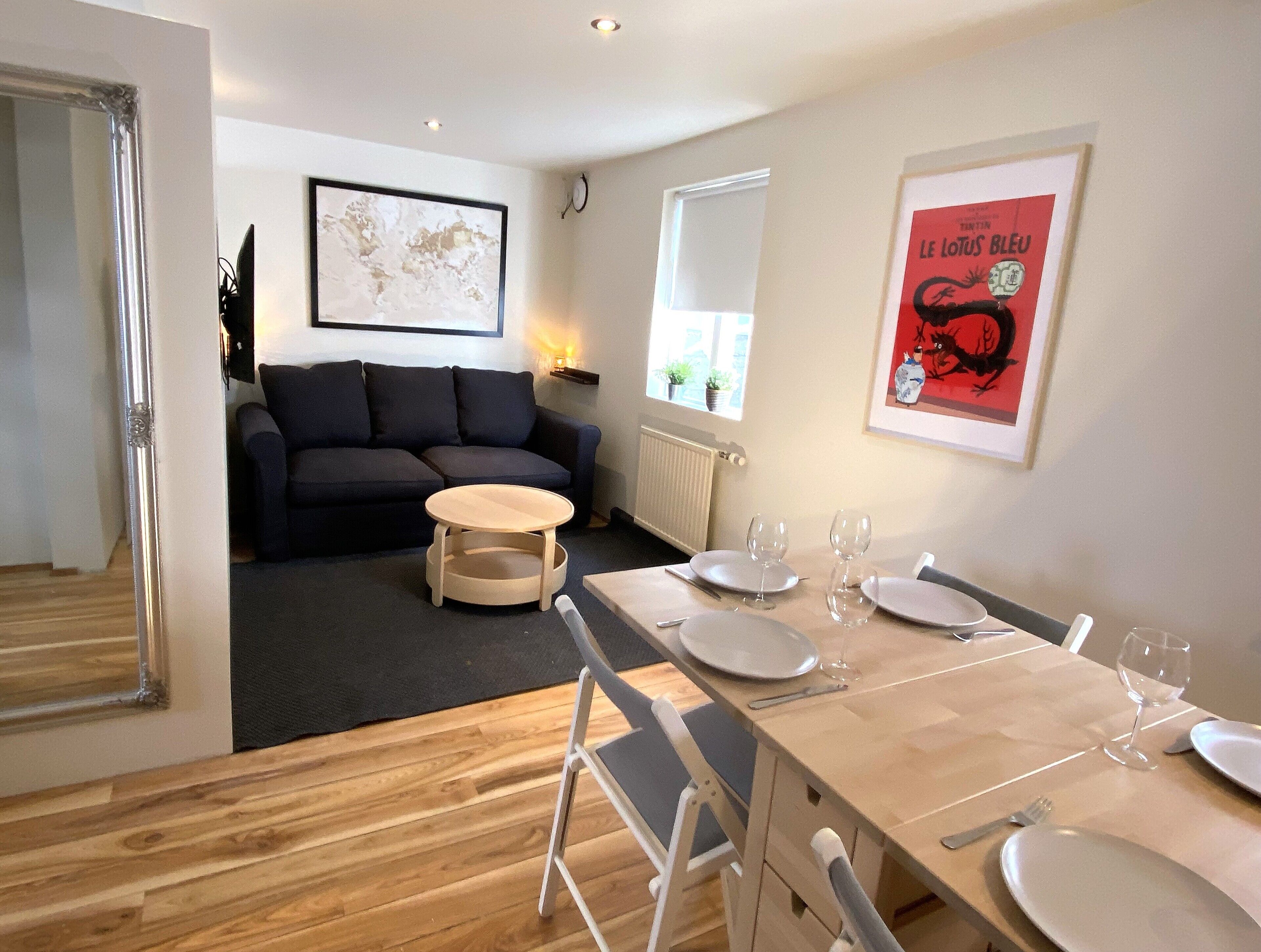 Un salon moderne avec un canapé foncé, une table basse ronde en bois et un coin repas avec une table en bois mise pour deux personnes.