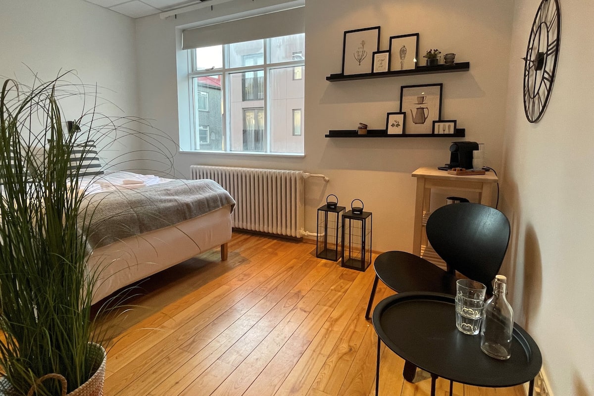 A modern room with a bed, a small table with a chair, a radiator, a window, and shelves with framed pictures.