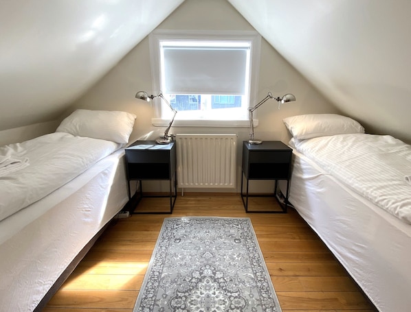 Un dormitorio abuhardillado pequeño y luminoso con dos camas individuales, un radiador y una ventana que deja entrar luz natural.