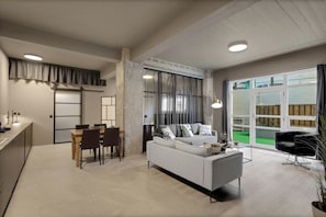 Ein modernes Wohnzimmer mit einer Couch, einem Esstisch und Stühlen. Ein großes Fenster mit Vorhängen, eine Deckenleuchte und Blick auf eine grüne Außenanlage.