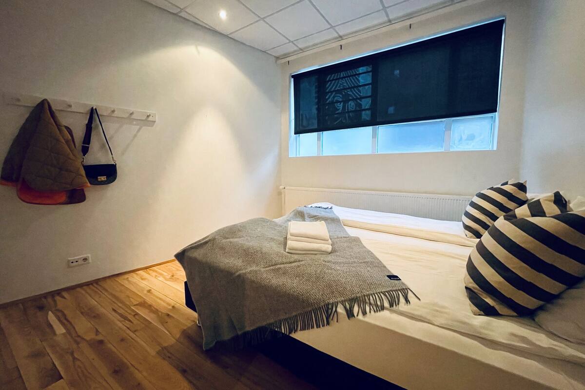 A bedroom with a bed, a window, a wall-mounted shelf, and a wooden floor.