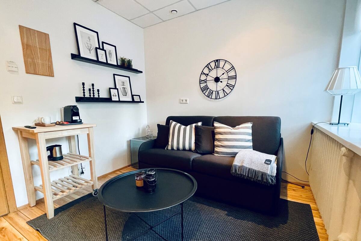A modern living room with a black sofa, a coffee table, a clock, and a shelf with framed pictures.