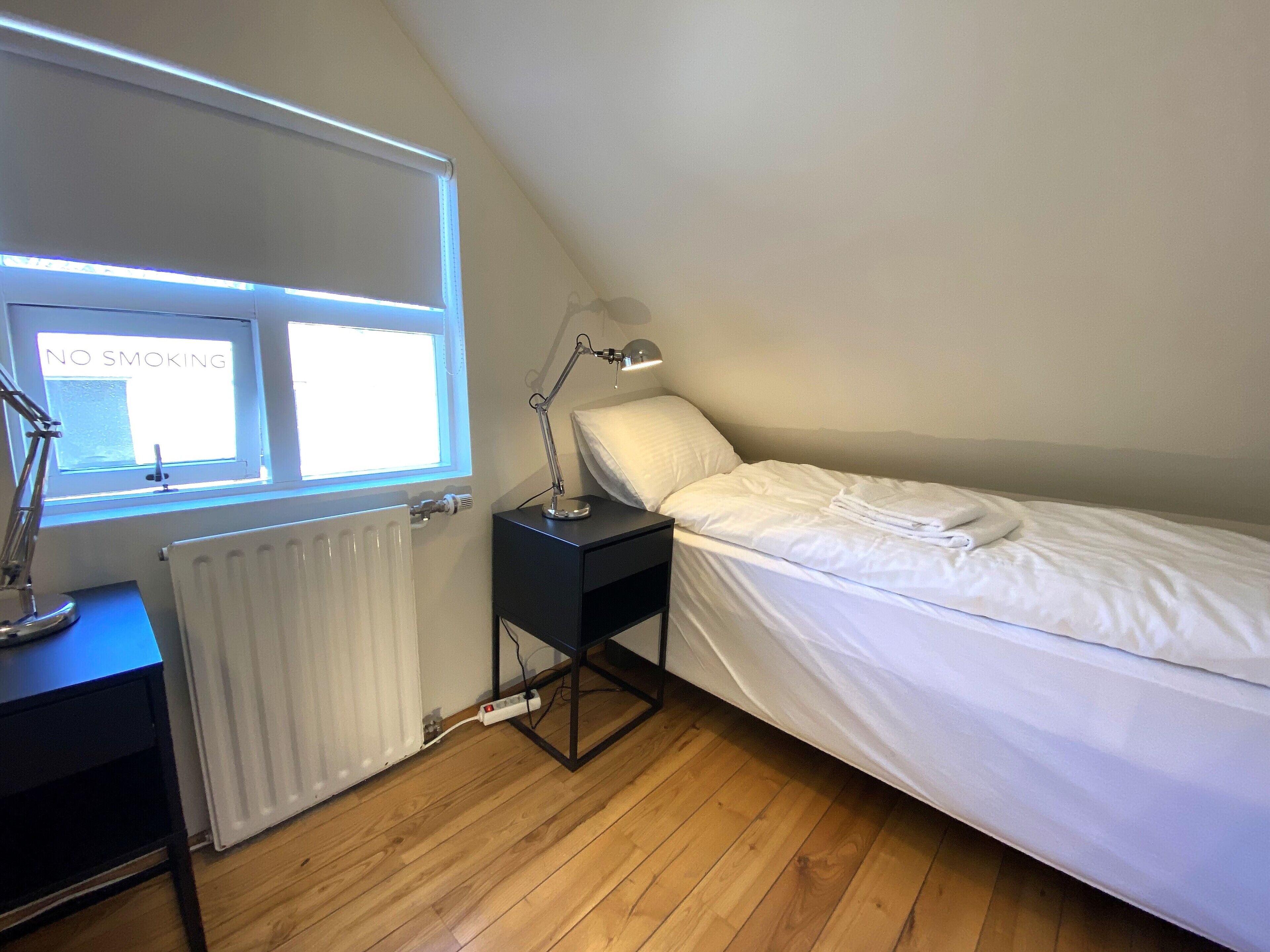 A bedroom with a single bed, a nightstand, a desk with a lamp, a radiator, and a window with a 'NO SMOKING' sign.