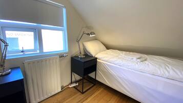 A bedroom with a single bed, a nightstand, a desk with a lamp, a radiator, and a window with a 'NO SMOKING' sign.