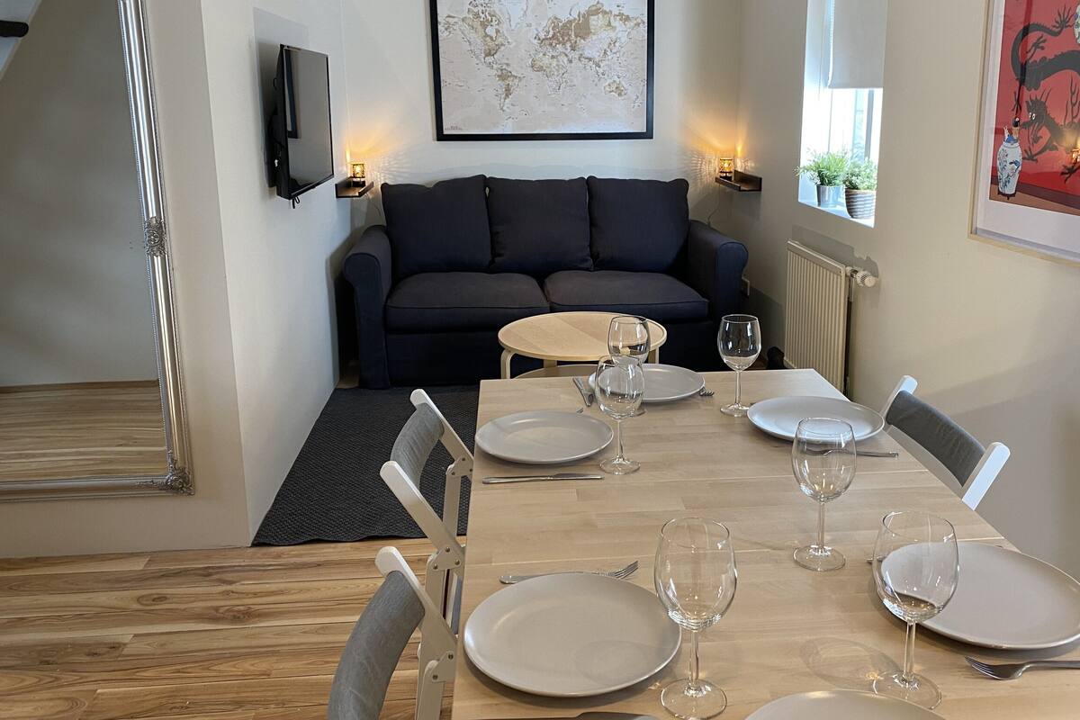 A dining area with a table set for four, a sofa, a TV, and a framed artwork on the wall.