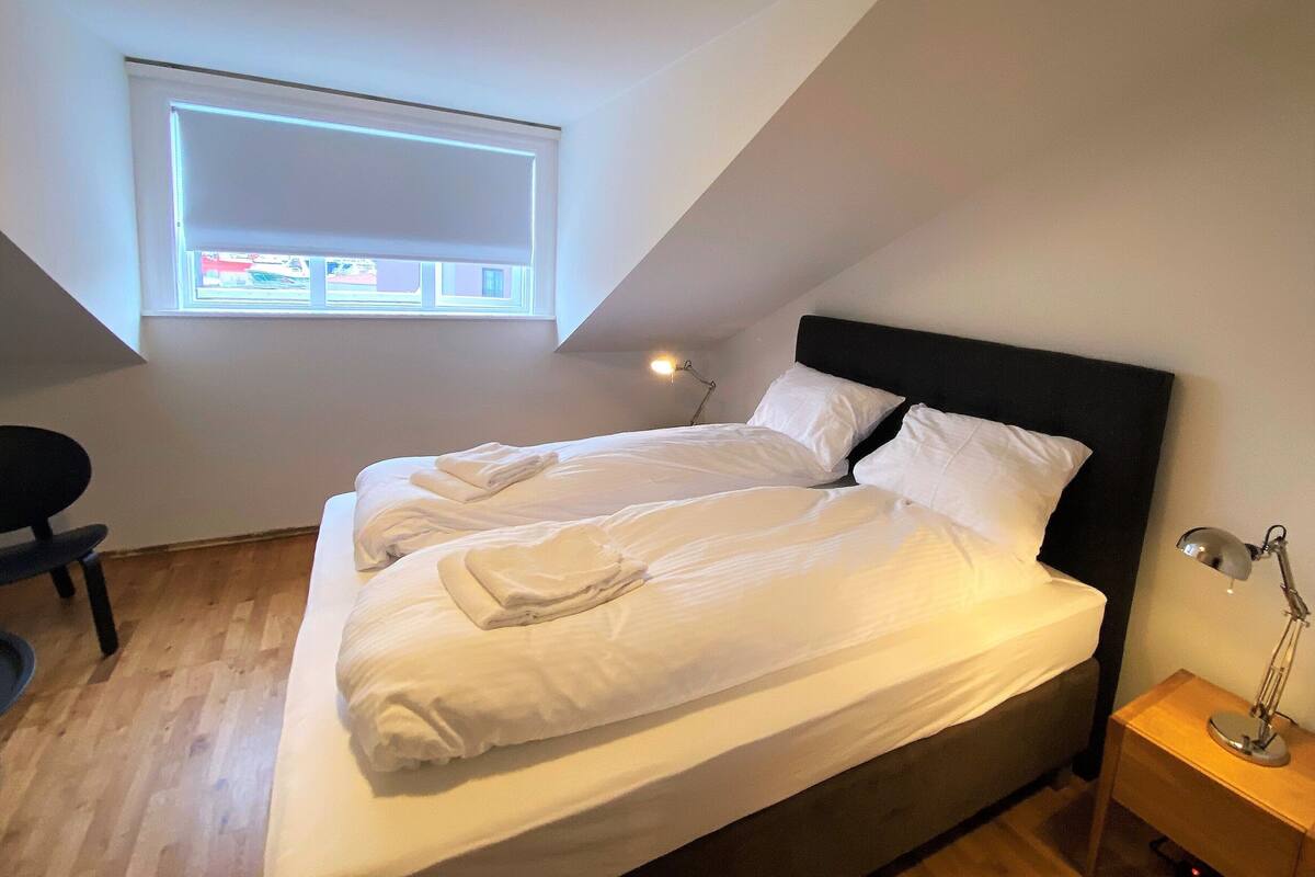 A bedroom with a bed, a desk, a chair, a window, and a nightstand with a lamp.