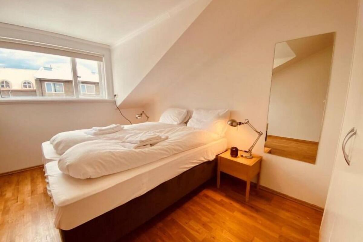 A bedroom with a bed, a nightstand, a lamp, a mirror, and a window.