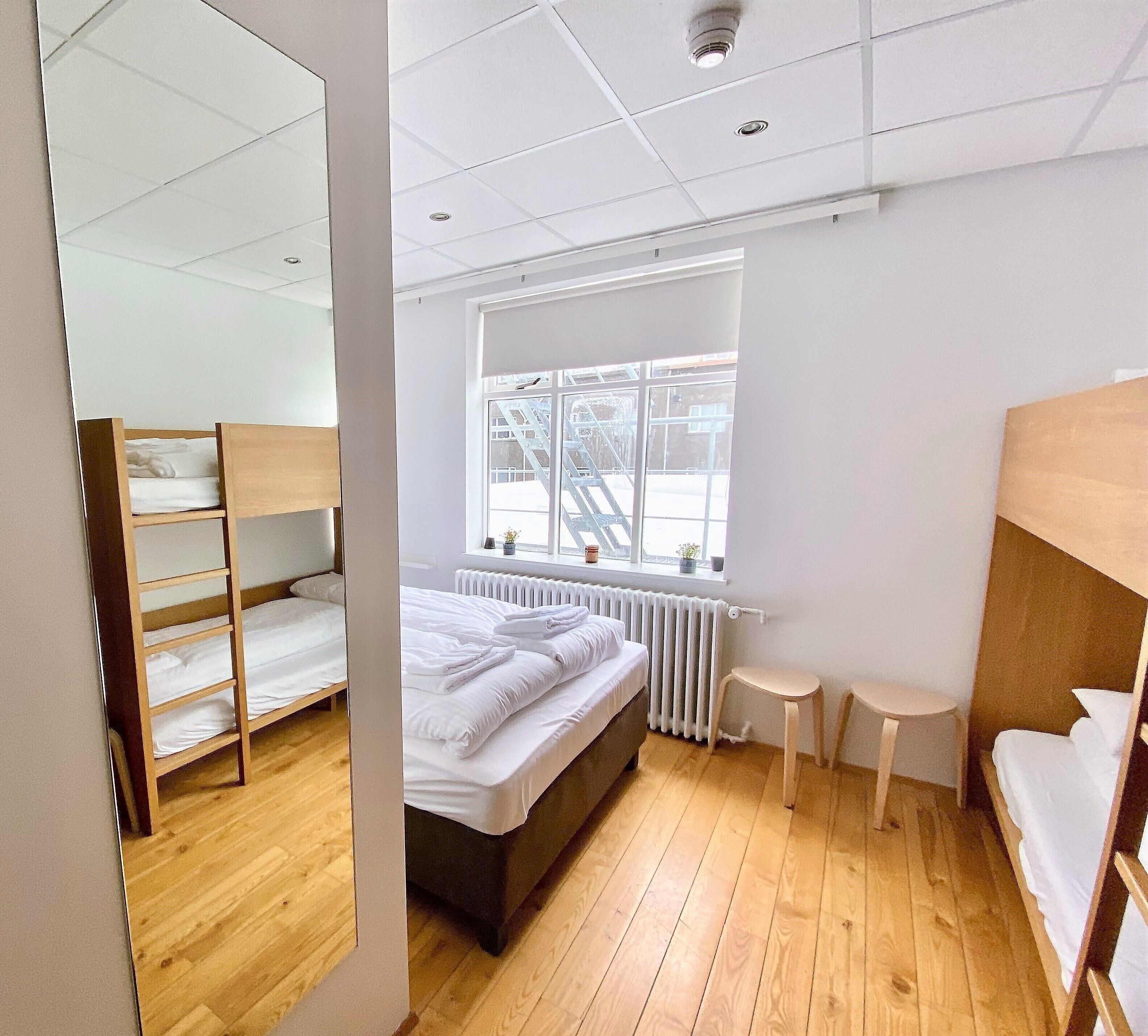 A dormitory room with bunk beds, a window with a view of a staircase, a radiator, and a small table.