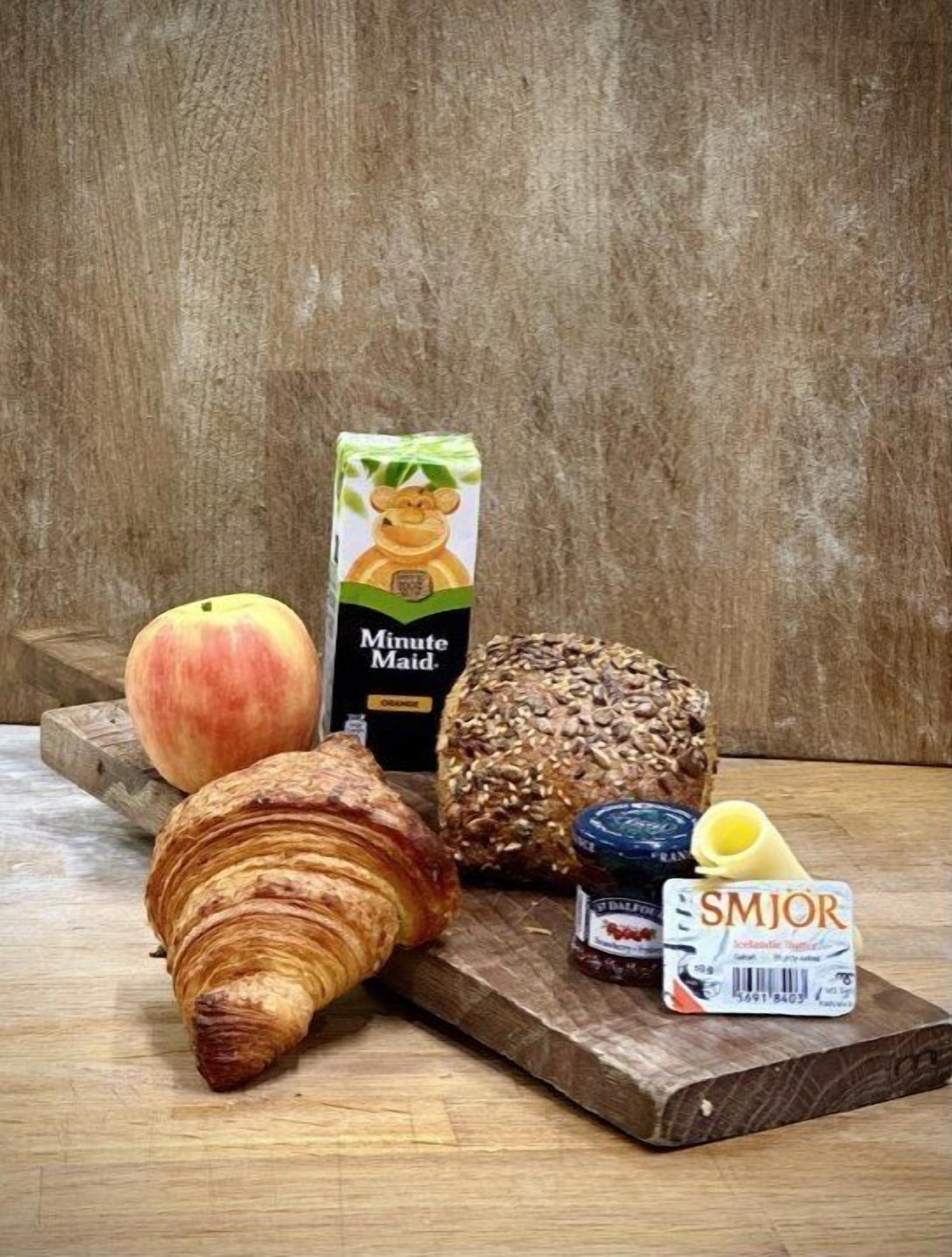 Una mesa de madera con un croissant, un melocotón, una hogaza de pan, un frasco de mermelada, un cartón de jugo de naranja Minute Maid y un paquete de helado SMJOR.