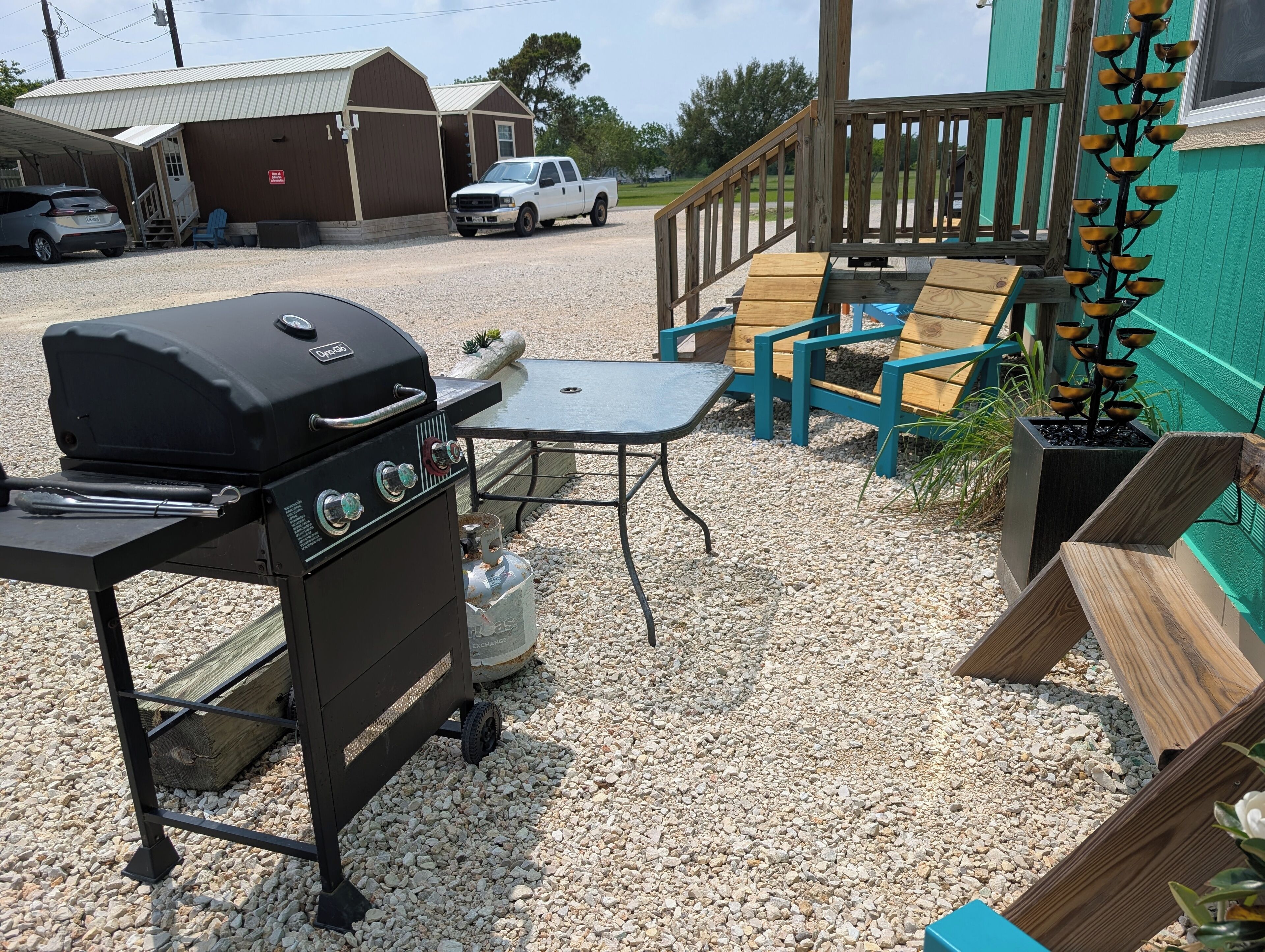 Grill, bord og udendørs møbler med træterrasse, og en varevogn parkeret i nærheden.