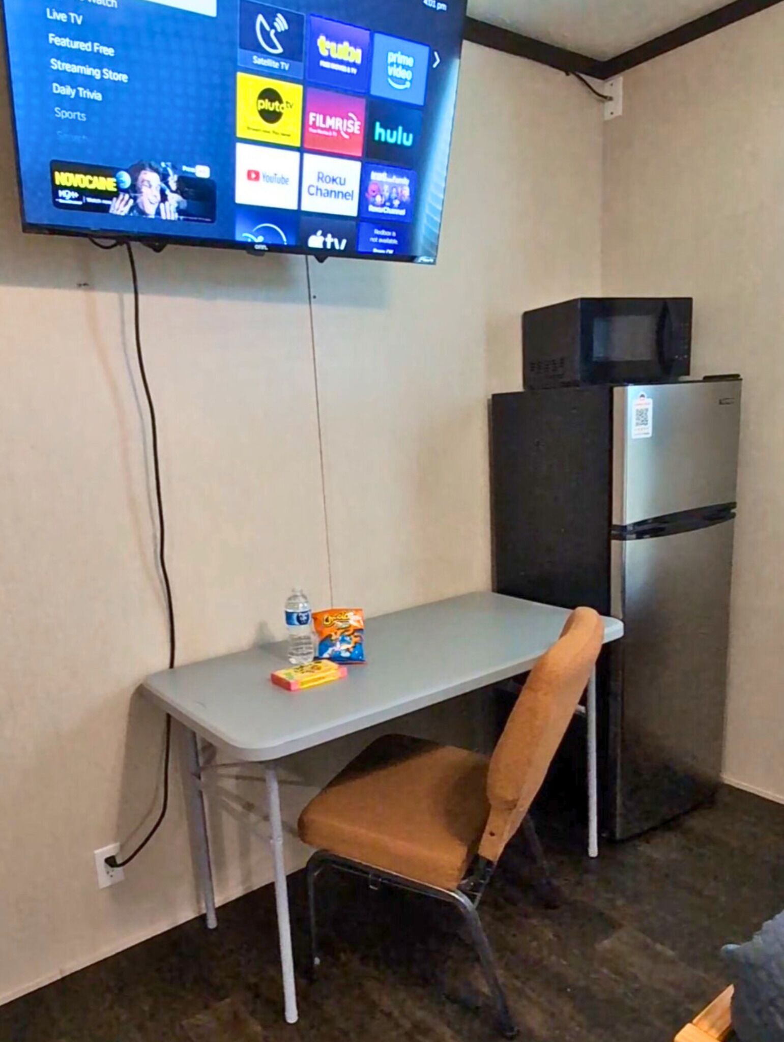 Una habitación pequeña con una televisión de pantalla plana mostrando diferentes servicios de streaming, un refrigerador, una mesa con una silla y una botella de agua.