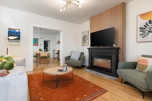 A modern living room with a fireplace, a flat-screen TV, and a dining area in the background.