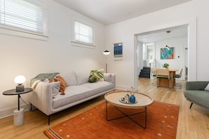 A modern living room with a grey sofa, a round coffee table, and a small side table with a lamp. There is a wooden dining table and chairs in the background.