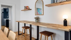 Une cuisine moderne avec un plan de travail en bois, deux tabourets, un miroir et une étagère contenant des livres et un vase.