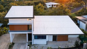 Ein modernes, zweistöckiges Haus mit Flachdach, umgeben von üppigem Grün und einem Sonnenuntergang im Hintergrund.