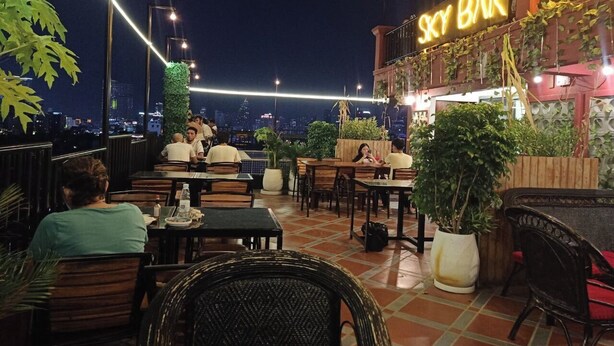 An outdoor seating area with tables and chairs, a neon sign for 'Sky Bar', and a city view in the background.