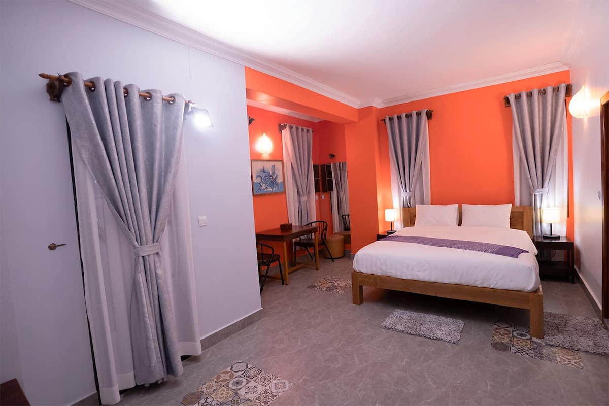 Ein Hotelzimmer mit einem Bett, einem Schreibtisch und einem Stuhl. Vor den Fenstern hängen Vorhänge und auf dem Schreibtisch steht eine Lampe.