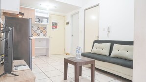 Ein modernes Wohnzimmer mit einer Couch, einem kleinen Tisch mit zwei Flaschen, einem Fernseher auf einem Schrank und einer Küchenzeile im Hintergrund.