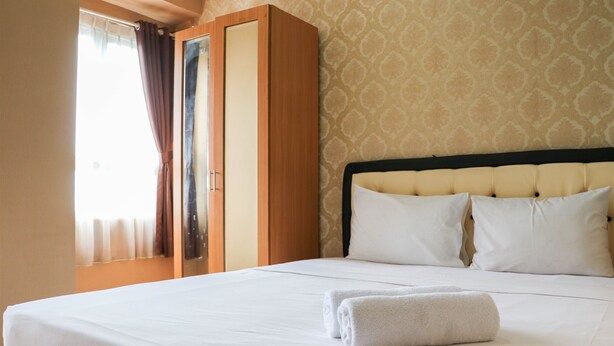A neatly made bed with white linens and a folded towel, a wooden wardrobe, and a patterned wallpaper. - Modern And Nice Studio At Titanium Square Apartment (Jakarta)