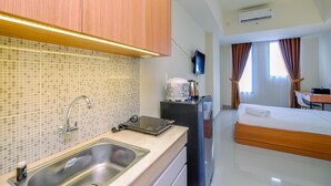 A compact hotel room with a kitchenette, a bed, a desk, and a TV. - Comfy Studio At Evenciio Margonda Apartment (Depok)