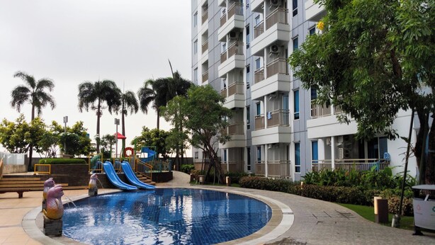 A swimming pool with slides, surrounded by a landscaped area with palm trees and a multi-story building in the background.
