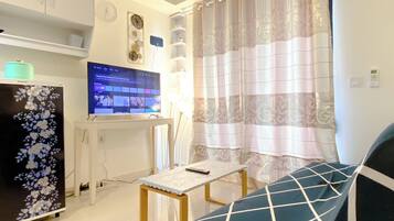 A living room with a flat-screen TV, a refrigerator, a table with a remote, and a patterned curtain.