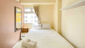 A hotel room with a bed, a nightstand, a mirror, a window with curtains, and a view of buildings outside.